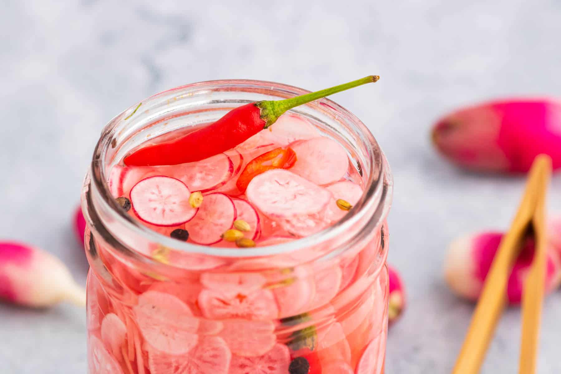 Quick Pickled Radishes couldn't be easier to make homemade. Have a jar at the ready in your refrigerator for burgers, hot dogs, sandwiches, tacos, salads and more. The top of an open glass jar filled with sliced radishes, red chili pepper, peppercorns, and coriander seeds.